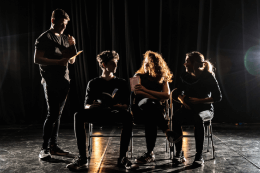 Three young people sit in a row with one person standing to the left of them.