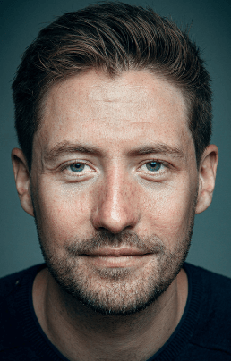 Headshot of white male with dark brown hair and stubble with a dark blue t-shirt on