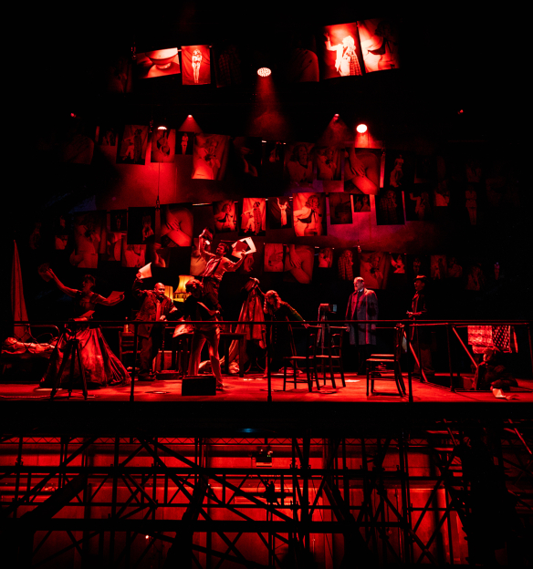 A dramatic stage scene lit in deep red tones, featuring multiple performers and props on a raised platform with hanging photographs or images in the background.