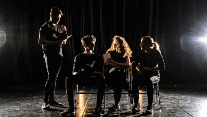 Three people sit in a row of chairs as one person stands at the end.