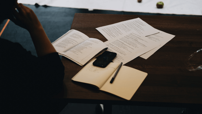 A silhouetted person leans their elbow on a table which holds scattered script pages, an open notebook, pen and mobile phone.