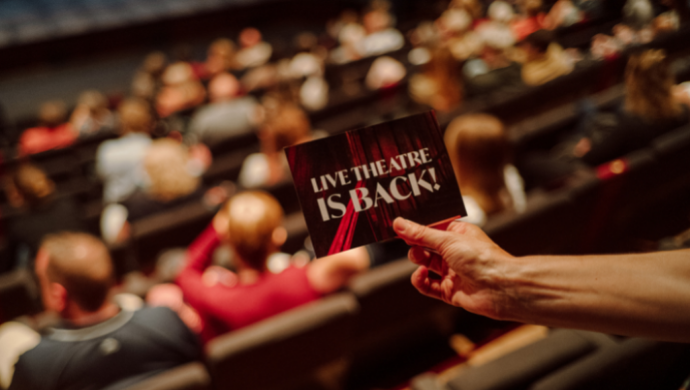 A close up of a card reading 'Live Theatre is Back!' being held in front of audience members sitting in the auditorium of the Main Stage.