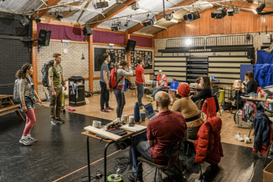 A group of actors rehearsing in a rehearsal space filled with props and tables, watched by six other people in various seats.