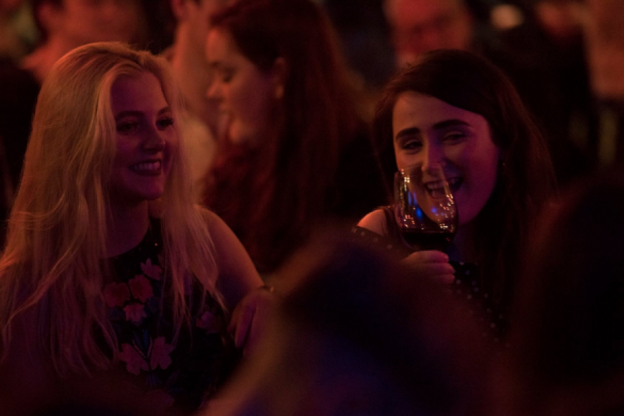 Two females sitting laughing, as one drinks from a wine glass.